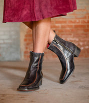 A person stands on a concrete floor wearing the Bed|Stü Bitsy black leather ankle boots, featuring a stacked heel and side zippers, paired with a burgundy skirt.