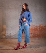 A woman stands indoors against a brick wall, hands in her pockets as she looks down. She wears a blue striped shirt, cropped jeans cinched with a brown belt, and finishes her look with the striking "Bitsy" red leather boots from Bed|Stü, featuring a stacked heel and square toe.
