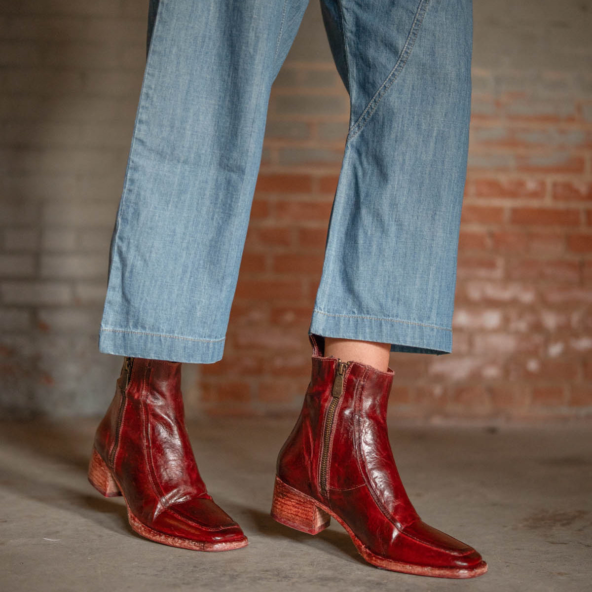 A person wearing ankle-length blue jeans and Bed|Stü "Bitsy" shiny red leather ankle boots with stacked heels and zippers, standing on a concrete floor in front of a brick wall.