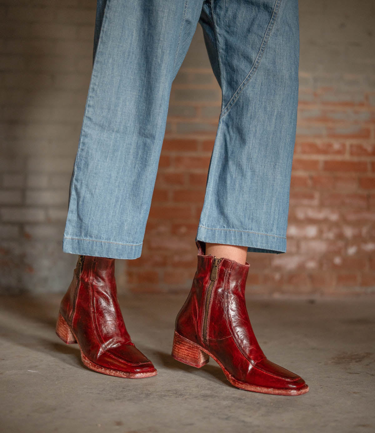 A person wearing ankle-length blue jeans and Bed|Stü "Bitsy" shiny red leather ankle boots with stacked heels and zippers, standing on a concrete floor in front of a brick wall.