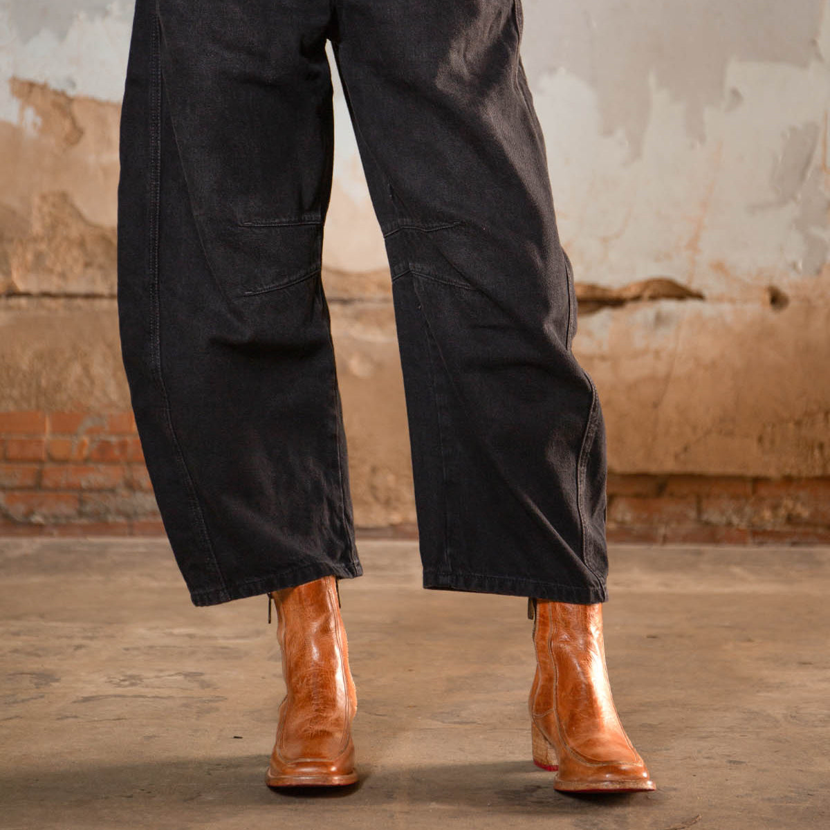 Wearing the Bed|Stü Bitsy brown leather ankle boots with a stacked heel and wide-leg black pants, the person stands on a worn concrete floor against a distressed wall backdrop.