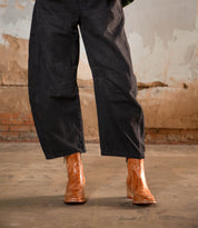 Wearing the Bed|Stü Bitsy brown leather ankle boots with a stacked heel and wide-leg black pants, the person stands on a worn concrete floor against a distressed wall backdrop.