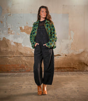 A woman stands indoors against a distressed wall, wearing black overalls, a green checked jacket, and the Bed|Stü Bitsy brown stacked heel leather boots, with her hands in her pockets and looking to the side.