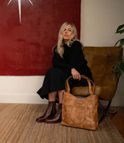 A woman with long blonde hair sits on a brown chair, wearing a black outfit and the Bed|Stü Bitsy brown boots with a stacked heel and square toe, while a large tan leather bag rests in front of her on a woven rug.
