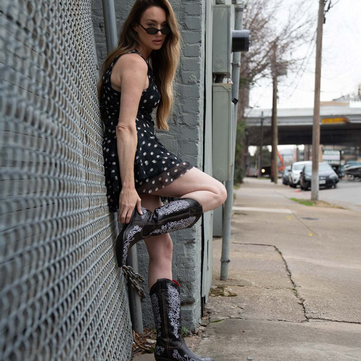 Wearing the Bed|Stü Blackjack Western tall boots adorned with floral embroidery, a person in a black polka dot dress and sunglasses leans against a gray brick wall by the sidewalk and street.