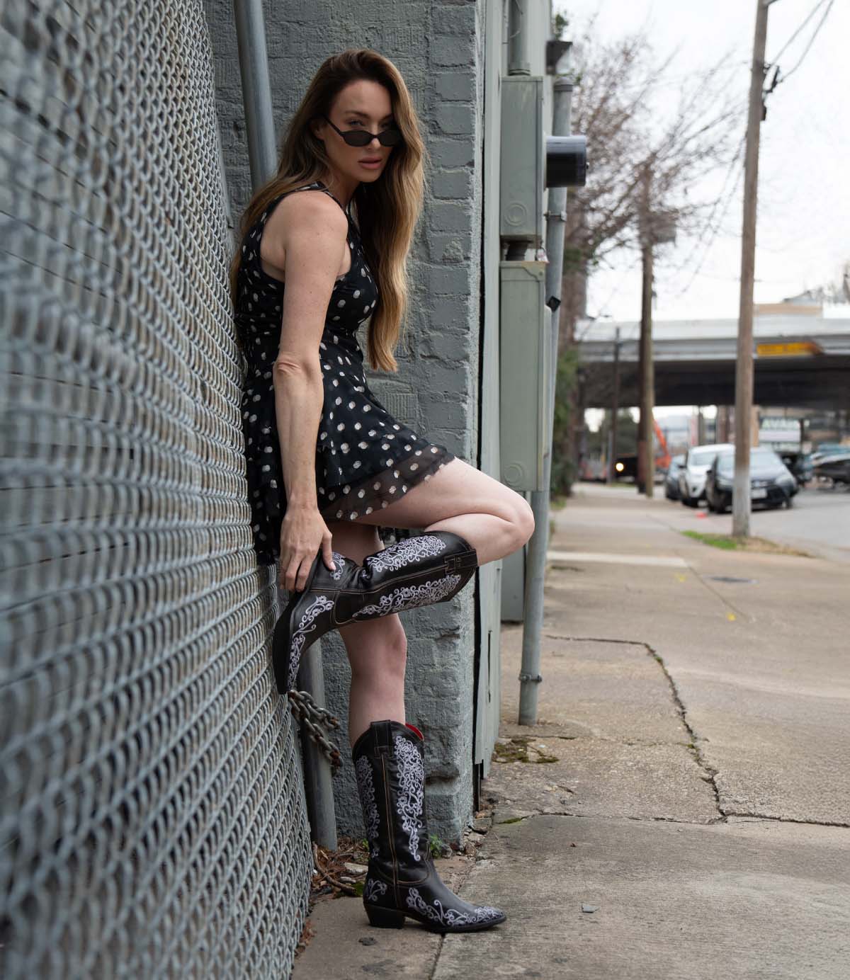 Wearing the Bed|Stü Blackjack Western tall boots adorned with floral embroidery, a person in a black polka dot dress and sunglasses leans against a gray brick wall by the sidewalk and street.