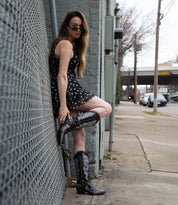 Wearing the Bed|Stü Blackjack Western tall boots adorned with floral embroidery, a person in a black polka dot dress and sunglasses leans against a gray brick wall by the sidewalk and street.