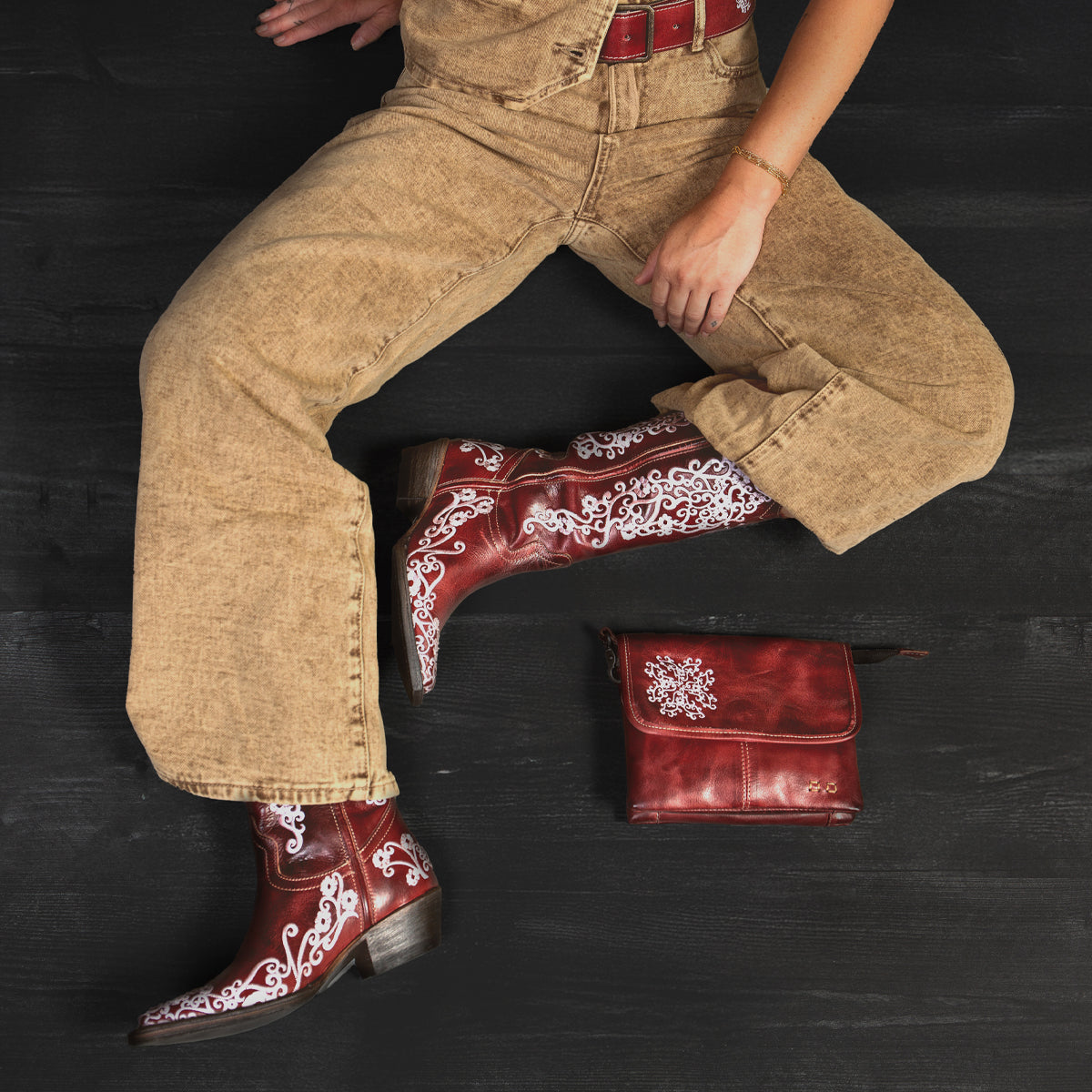 A person in a tan jumpsuit and striking Bed|Stü Blackjack red embroidered cowboy boots with a snip toe sits cross-legged on a black surface, with a matching red leather bag placed nearby.