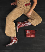 A person in a tan jumpsuit and striking Bed|Stü Blackjack red embroidered cowboy boots with a snip toe sits cross-legged on a black surface, with a matching red leather bag placed nearby.