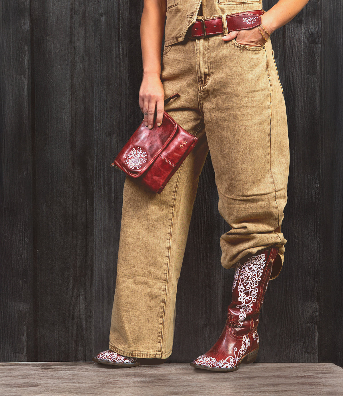 A person stands against a dark wooden background wearing tan pants, holding a red clutch, and showcasing the Bed|Stü Blackjack Western tall boots featuring red and white floral embroidery and a classic snip toe.