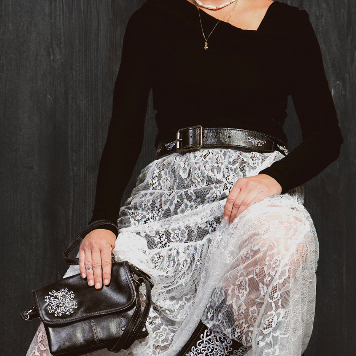 A person sits against a dark background, wearing a black top, a white lace skirt, and a black belt with an antique-finished buckle. Necklaces are layered across their chest as they hold the Bed|Stü Blaze JB purse—an elegant black bag featuring intricate embroidered scrollwork. Only the lower face and body are visible.