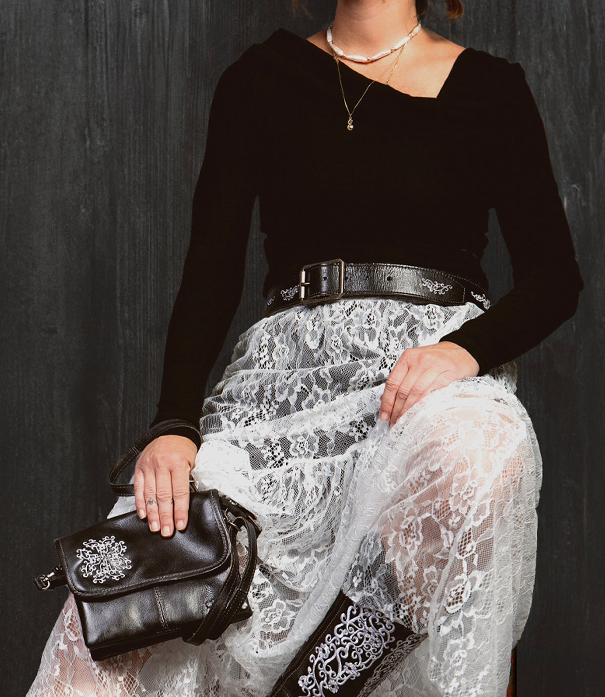 A person sits against a dark background, wearing a black top, a white lace skirt, and a black belt with an antique-finished buckle. Necklaces are layered across their chest as they hold the Bed|Stü Blaze JB purse—an elegant black bag featuring intricate embroidered scrollwork. Only the lower face and body are visible.