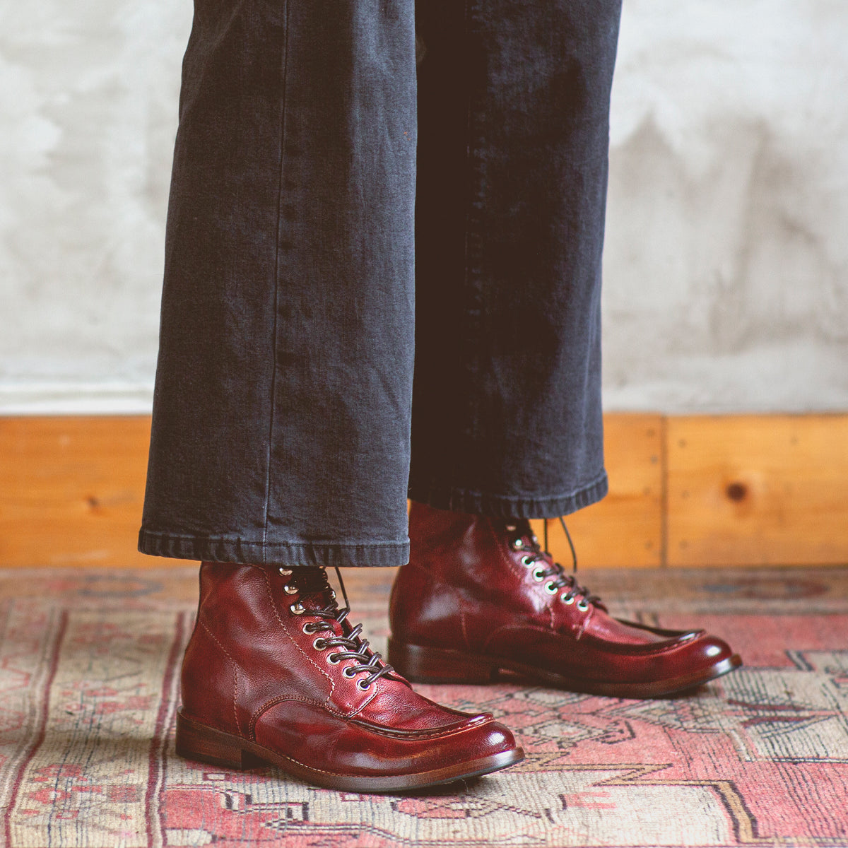 A person wearing dark pants and red Blimey boots from Bed|Stü stands on a patterned rug near a light-colored wall with wood trim. These full-grain leather boat shoe boots, handcrafted in Italy, exude both timeless style and exceptional craftsmanship.