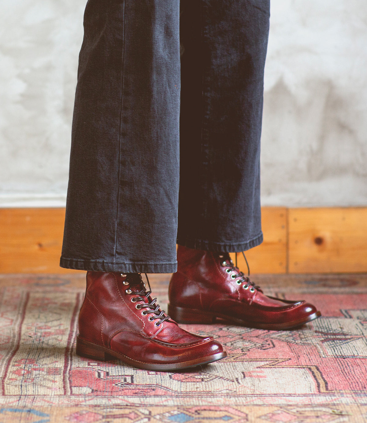 A person wearing dark pants and red Blimey boots from Bed|Stü stands on a patterned rug near a light-colored wall with wood trim. These full-grain leather boat shoe boots, handcrafted in Italy, exude both timeless style and exceptional craftsmanship.