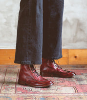 A person wearing dark pants and red Blimey boots from Bed|Stü stands on a patterned rug near a light-colored wall with wood trim. These full-grain leather boat shoe boots, handcrafted in Italy, exude both timeless style and exceptional craftsmanship.