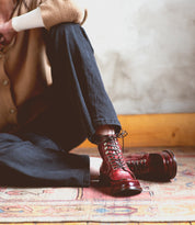 Person seated on a patterned rug, wearing dark pants, a tan long-sleeve shirt, and Bed|Stü Blimey shiny red full-grain leather boots handcrafted in Italy, with one knee bent and their face out of frame.