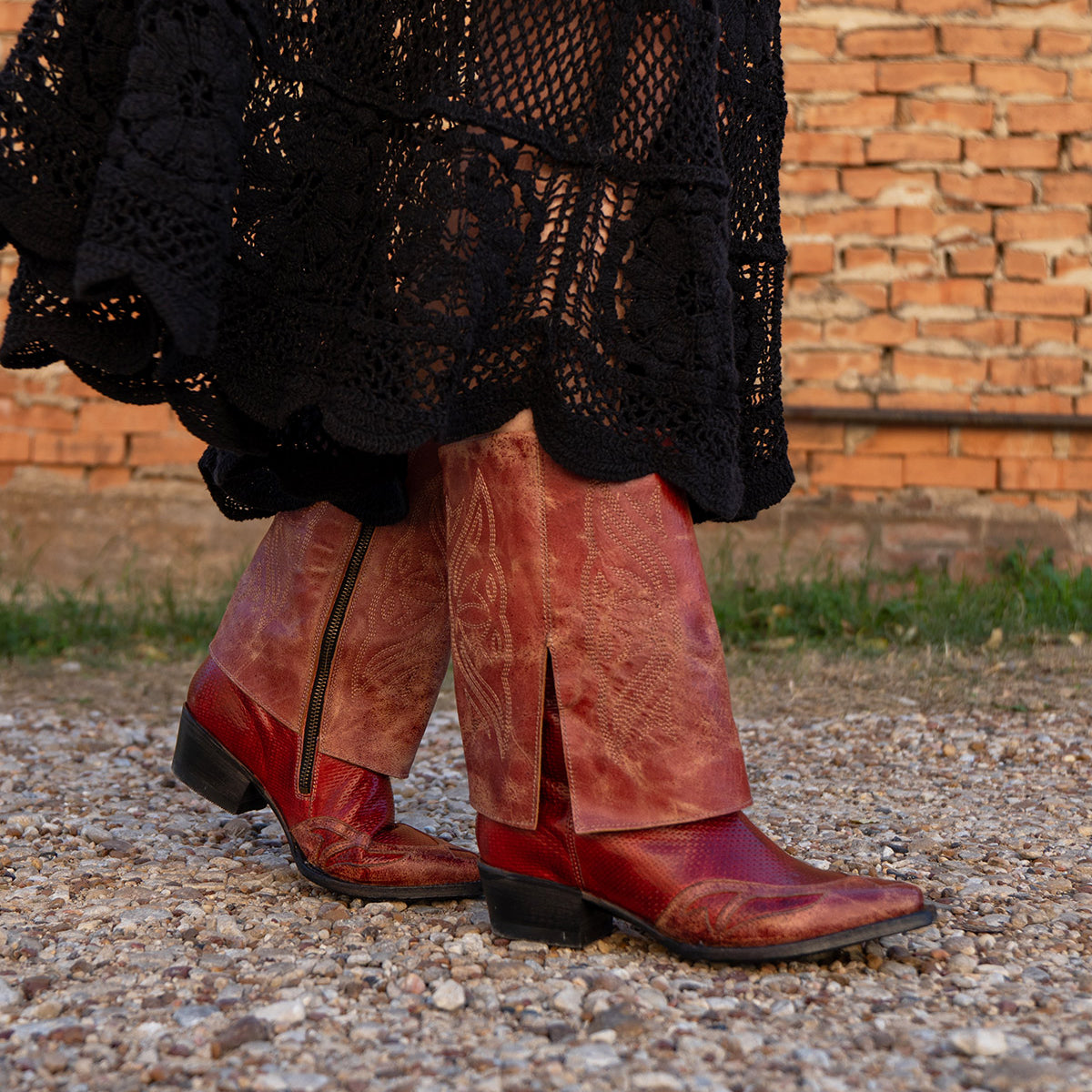 Wearing the Blind handcrafted western boots by Bed|Stü and a long black crocheted skirt, a person walks on a gravel path near a brick wall.