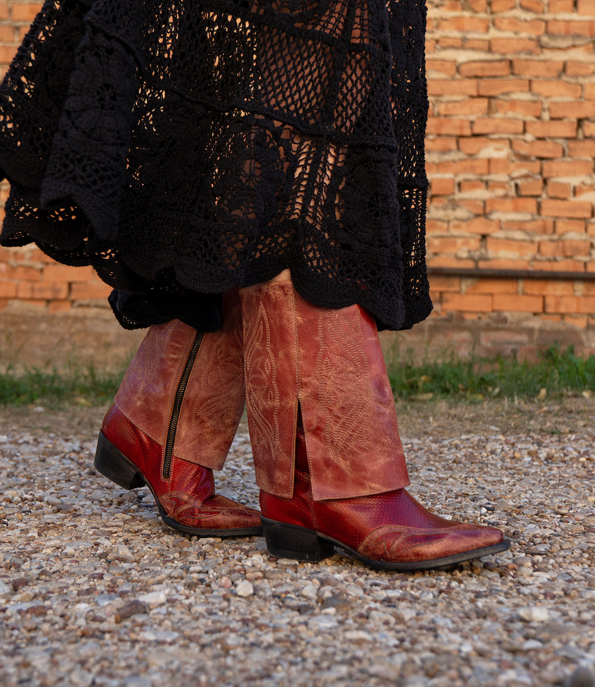 Wearing the Blind handcrafted western boots by Bed|Stü and a long black crocheted skirt, a person walks on a gravel path near a brick wall.