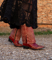 Wearing the Blind handcrafted western boots by Bed|Stü and a long black crocheted skirt, a person walks on a gravel path near a brick wall.