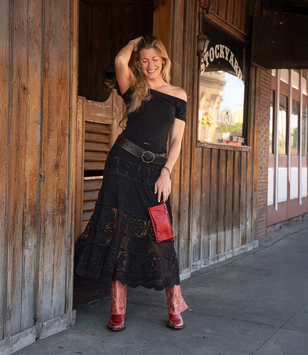 A woman in a black top and skirt, wearing Bed|Stü’s handcrafted Blind western boots in vibrant red and holding a matching red clutch, stands smiling by a wooden building with a "Stockyards" sign in the background.