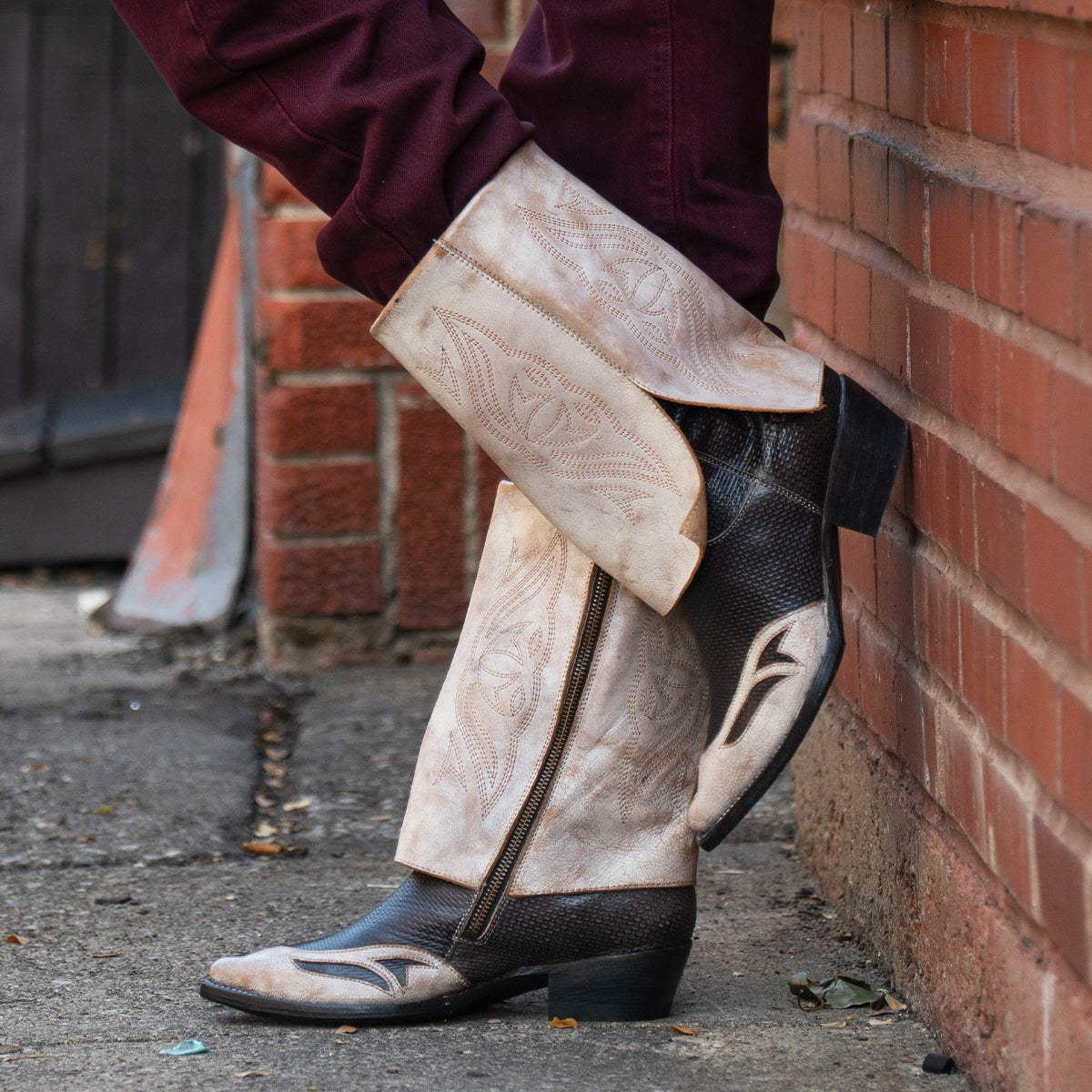 A person wearing the Bed|Stü Blind handcrafted western boots in tan and black with maroon pants, leaning against a brick wall on a city sidewalk.