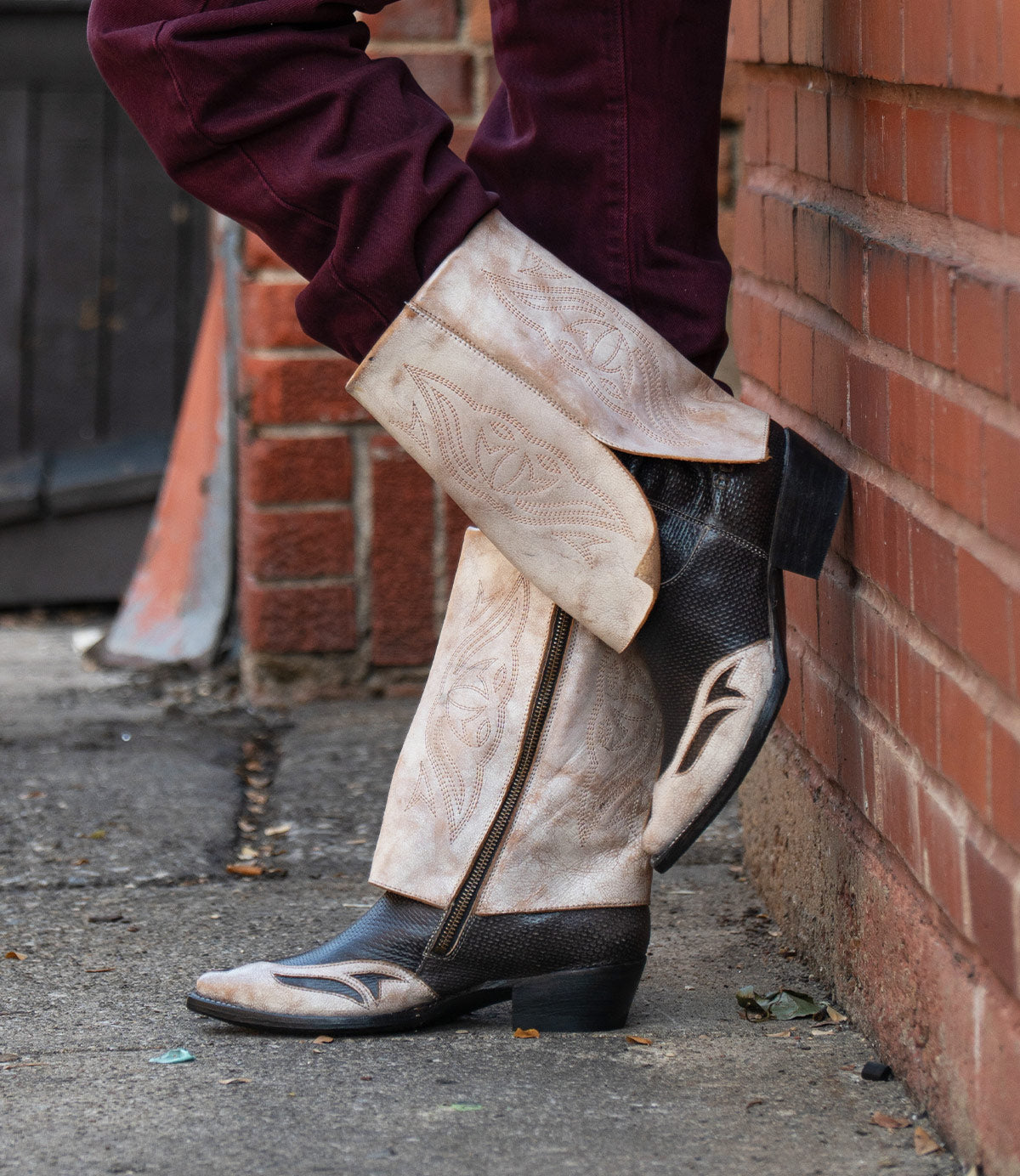 A person wearing the Bed|Stü Blind handcrafted western boots in tan and black with maroon pants, leaning against a brick wall on a city sidewalk.