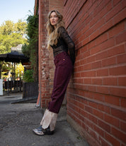 A woman in a black lace top and burgundy jeans leans against a brick wall outdoors, wearing Bed|Stü's Blind handcrafted western boots in a stylish two-tone design.