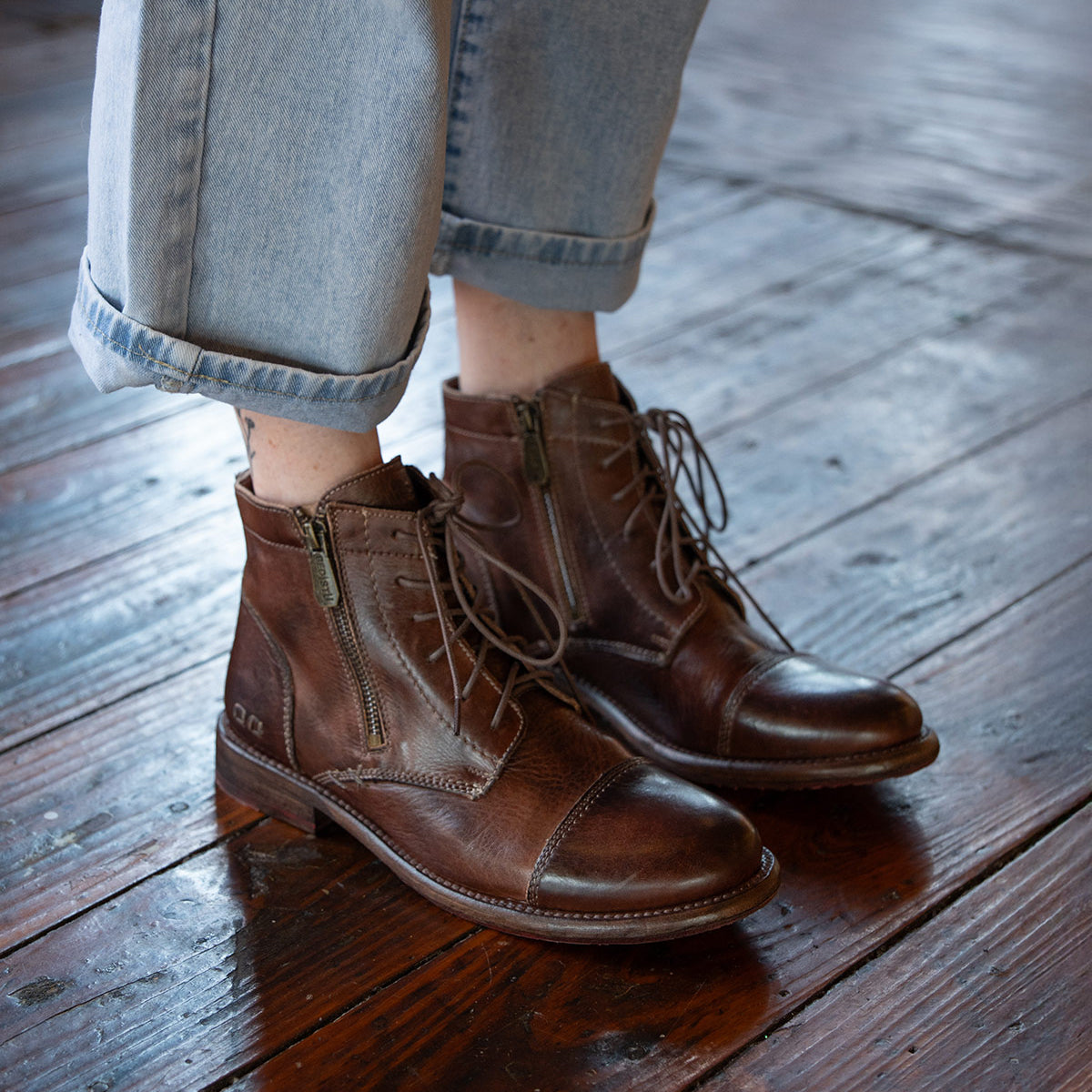 Person wearing rolled-up light blue jeans and Bed|Stü Bonnie II brown leather cap toe lace-up ankle boots, featuring a Goodyear-welted outsole, standing on a polished wooden floor.