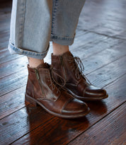 Person wearing rolled-up light blue jeans and Bed|Stü Bonnie II brown leather cap toe lace-up ankle boots, featuring a Goodyear-welted outsole, standing on a polished wooden floor.