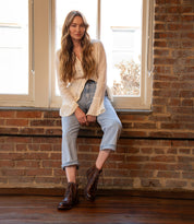 A person with long hair in a white shirt, light blue jeans, and the Bed|Stü Bonnie II brown combat ankle boots with a Goodyear-welted outsole sits on a windowsill against an exposed brick wall, looking at the camera.