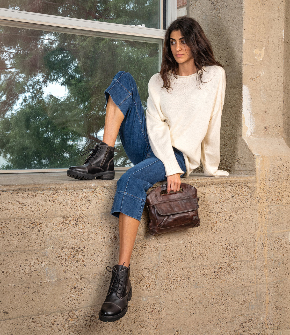 A woman wearing a cream sweater and cropped jeans sits on a ledge, wearing dark leather Bed|Stü Bonnie IV combat boots with dual side zippers and holding a brown leather handbag.