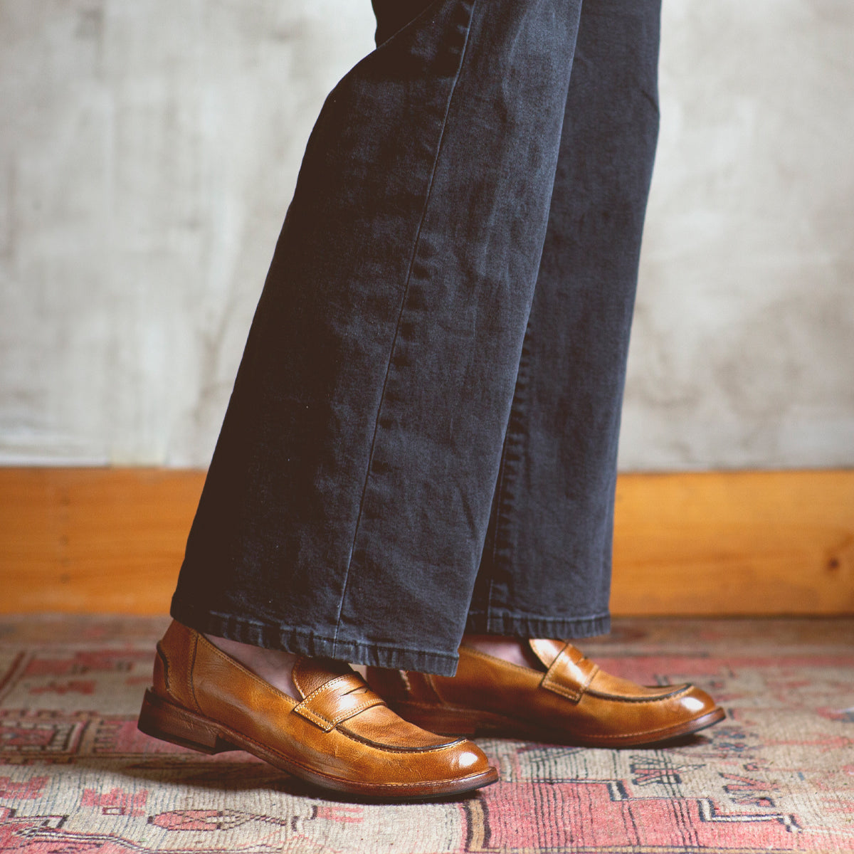 A person wearing black pants and classic style Bed|Stü Bonus brown Italian leather penny loafers stands on a patterned rug with a wooden baseboard and gray textured wall in the background.