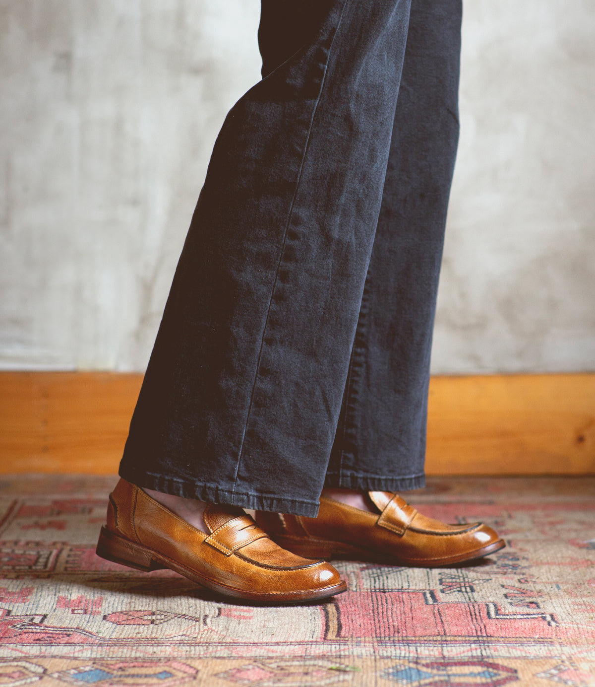 A person wearing black pants and classic style Bed|Stü Bonus brown Italian leather penny loafers stands on a patterned rug with a wooden baseboard and gray textured wall in the background.