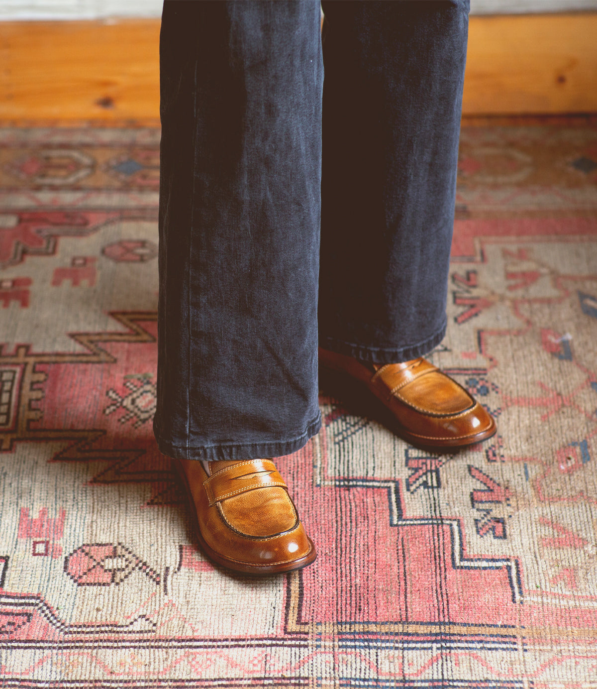 Wearing dark pants, the person showcases a classic style standing on a patterned rug with geometric designs, featuring the Bonus brown Italian leather penny loafers by Bed|Stü.