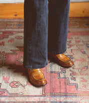 Wearing dark pants, the person showcases a classic style standing on a patterned rug with geometric designs, featuring the Bonus brown Italian leather penny loafers by Bed|Stü.