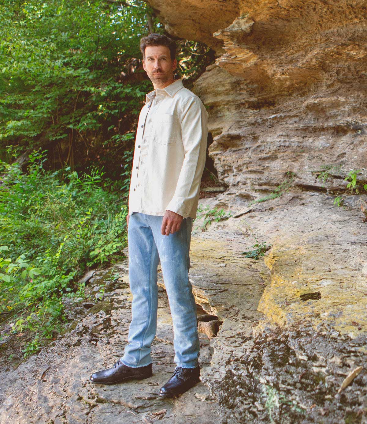 A man in a light shirt and jeans stands on a rocky surface outdoors, showcasing the Bradley boots by Bed|Stü—a timeless pair of classic lace-up footwear—surrounded by greenery and a striking rock formation in the background.