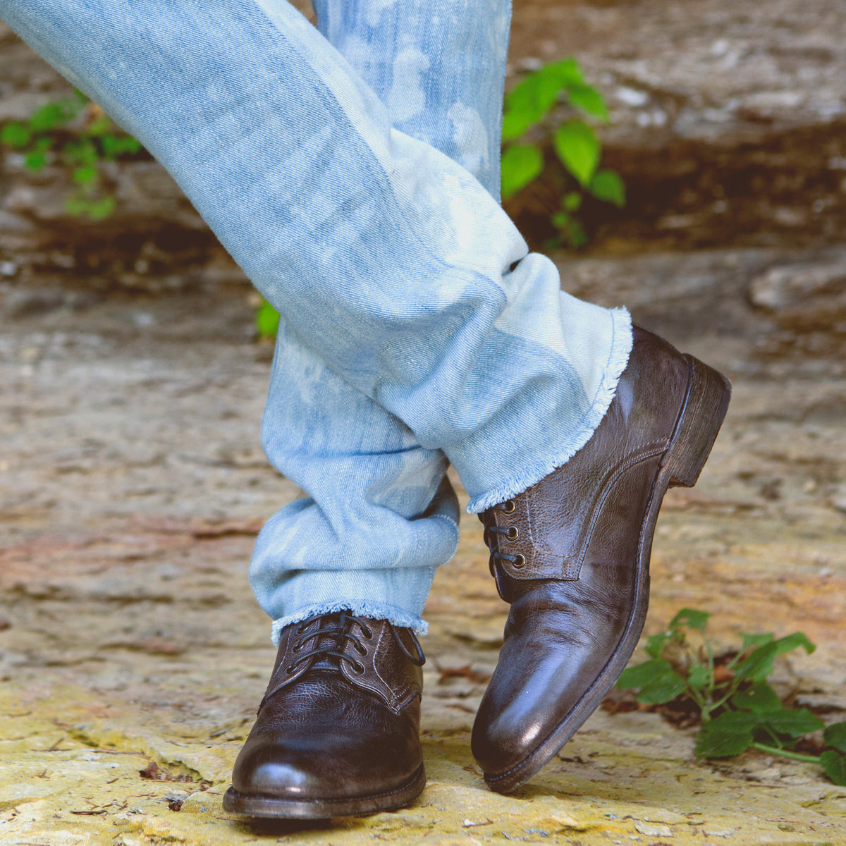 Dressed in light blue jeans and the Bradley black leather lace-up boots by Bed|Stü, a person stands outdoors on a stone surface with one leg crossed over the other, showcasing effortless everyday style.