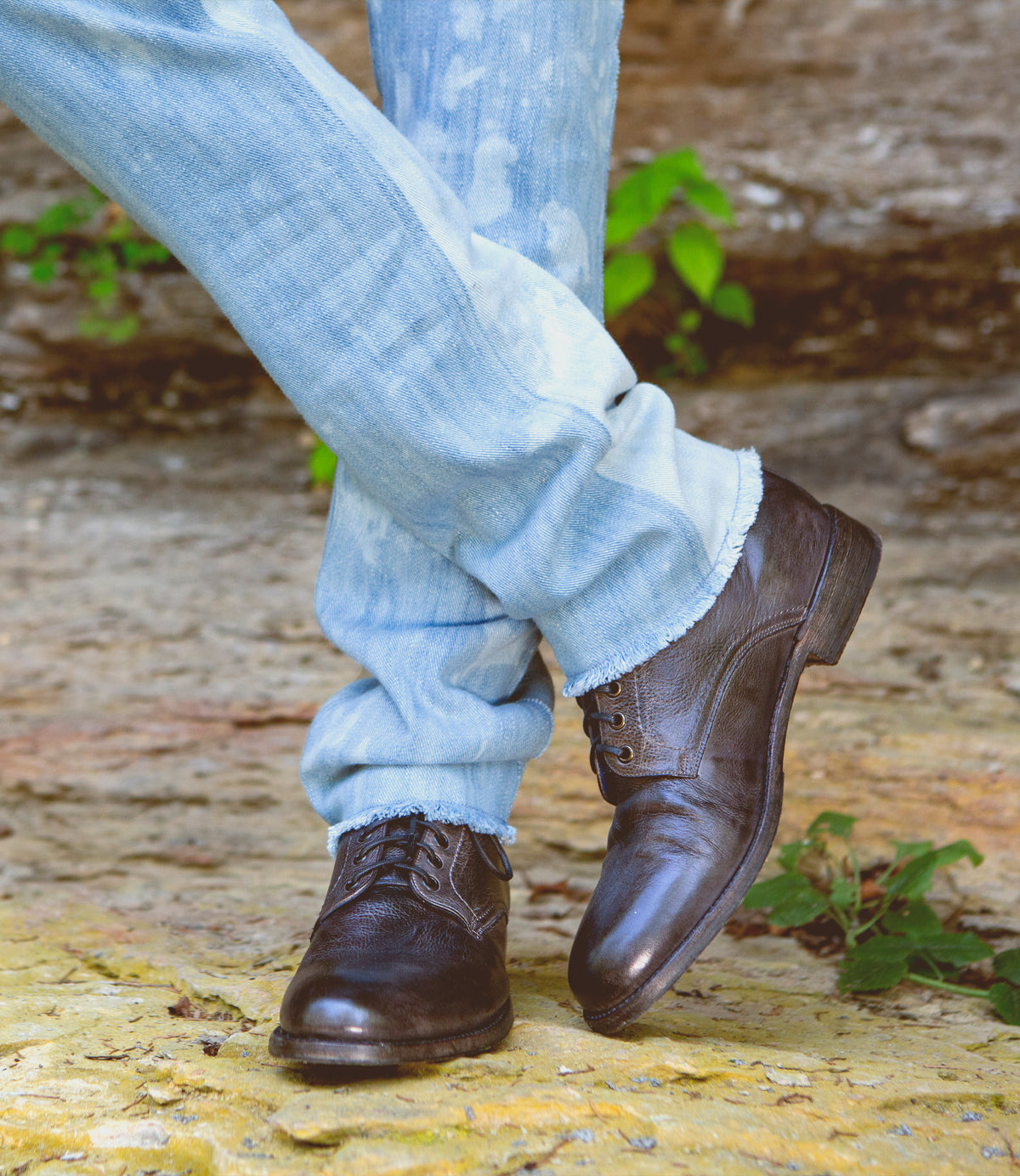 Dressed in light blue jeans and the Bradley black leather lace-up boots by Bed|Stü, a person stands outdoors on a stone surface with one leg crossed over the other, showcasing effortless everyday style.