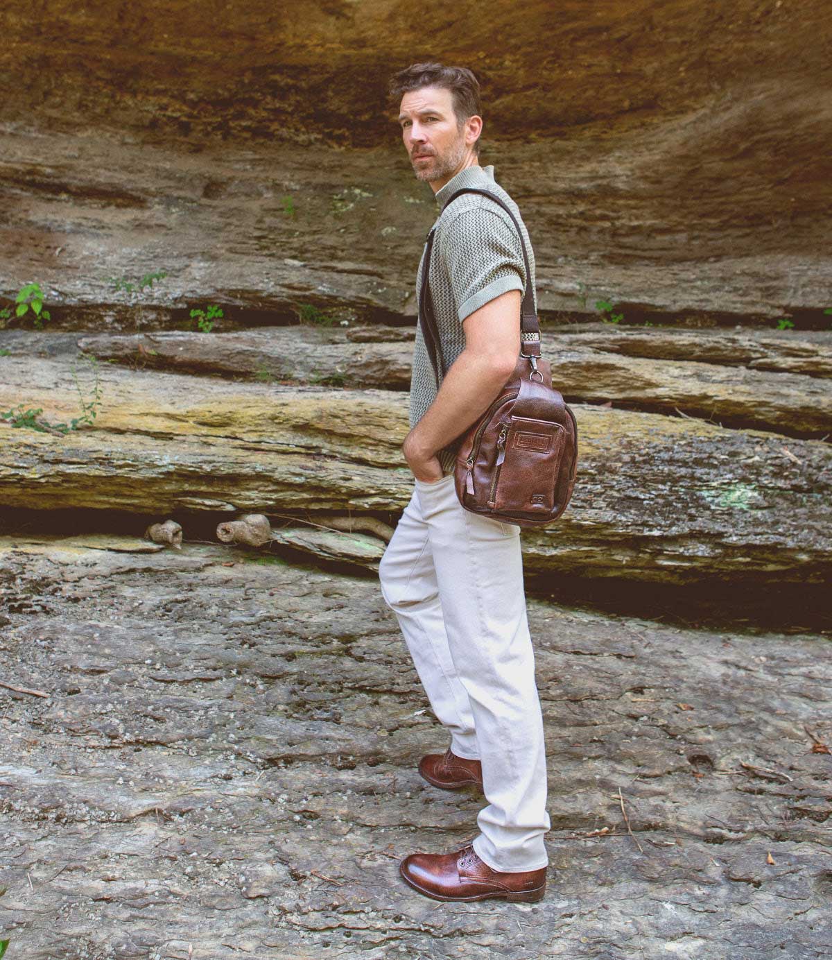 A man stands on rocky ground near a stone wall, wearing a green shirt, white pants, and timeless Bed|Stü Bradley brown lace-up boots, with a brown leather bag over his shoulder.