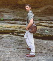 A man stands on rocky ground near a stone wall, wearing a green shirt, white pants, and timeless Bed|Stü Bradley brown lace-up boots, with a brown leather bag over his shoulder.