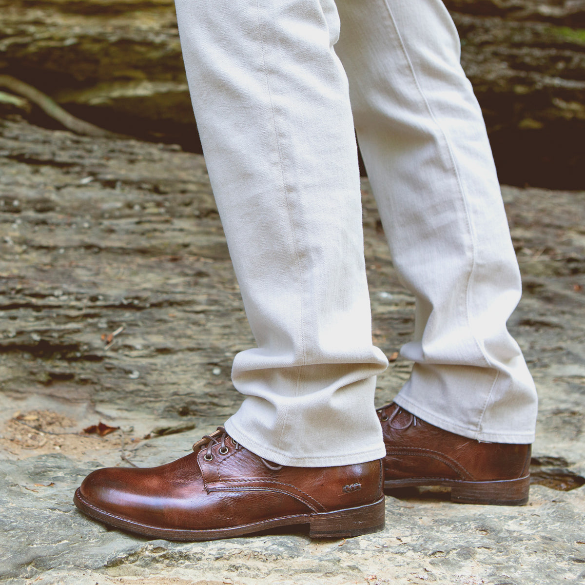 A person wearing light-colored pants and the timeless Bradley boots from Bed|Stü—crafted from brown leather with classic lace-up detailing—stands on a rocky outdoor surface, showcasing their versatility for everyday wear.