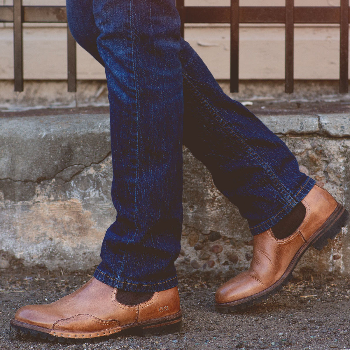 A person wearing blue jeans and Bed|Stü’s Brady Trek brown leather Chelsea boots with a treaded outsole stands with one foot crossed in front of the other on a concrete sidewalk.