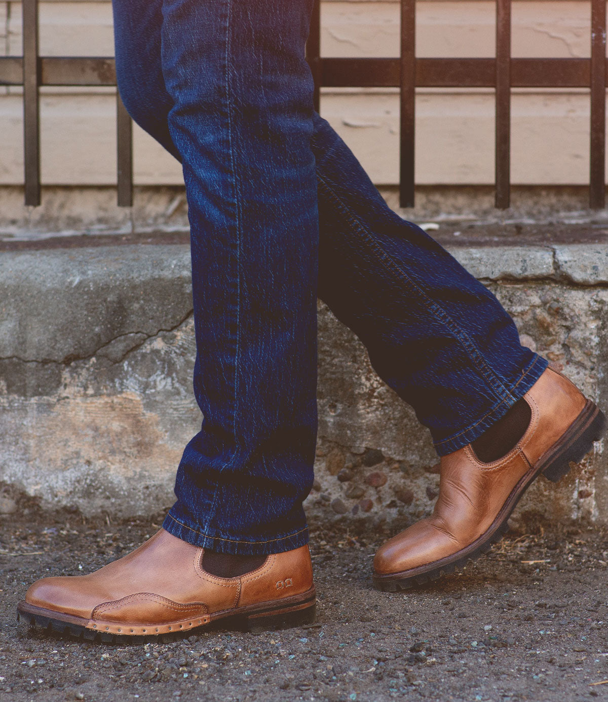 A person wearing blue jeans and Bed|Stü’s Brady Trek brown leather Chelsea boots with a treaded outsole stands with one foot crossed in front of the other on a concrete sidewalk.