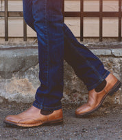 A person wearing blue jeans and Bed|Stü’s Brady Trek brown leather Chelsea boots with a treaded outsole stands with one foot crossed in front of the other on a concrete sidewalk.
