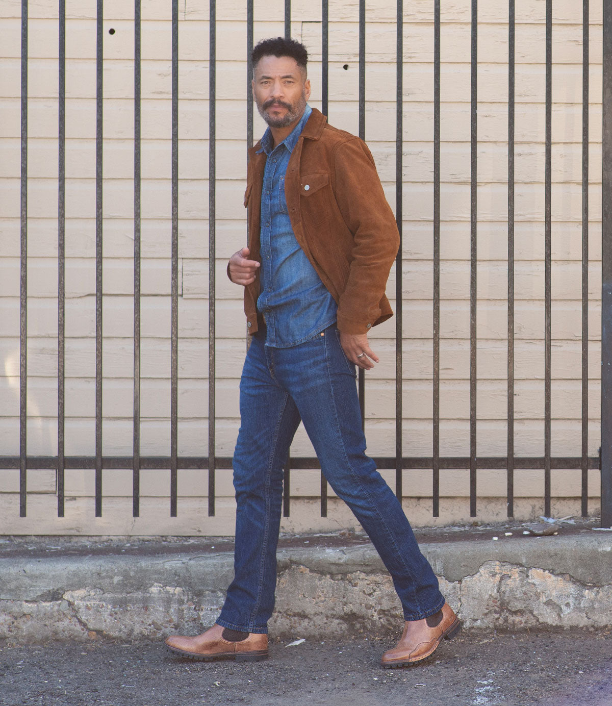 A man walks outdoors in front of a wooden wall with metal bars, wearing a brown suede jacket, denim shirt, blue jeans, and Bed|Stü Brady Trek classic Chelsea boots with a treaded outsole.