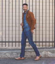 A man walks outdoors in front of a wooden wall with metal bars, wearing a brown suede jacket, denim shirt, blue jeans, and Bed|Stü Brady Trek classic Chelsea boots with a treaded outsole.