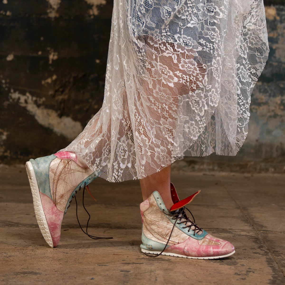 A person wearing the Bed|Stü Breath pastel-colored high-top statement sneakers and a sheer white lace dress stands on a worn concrete floor, with a textured, weathered wall in the background.