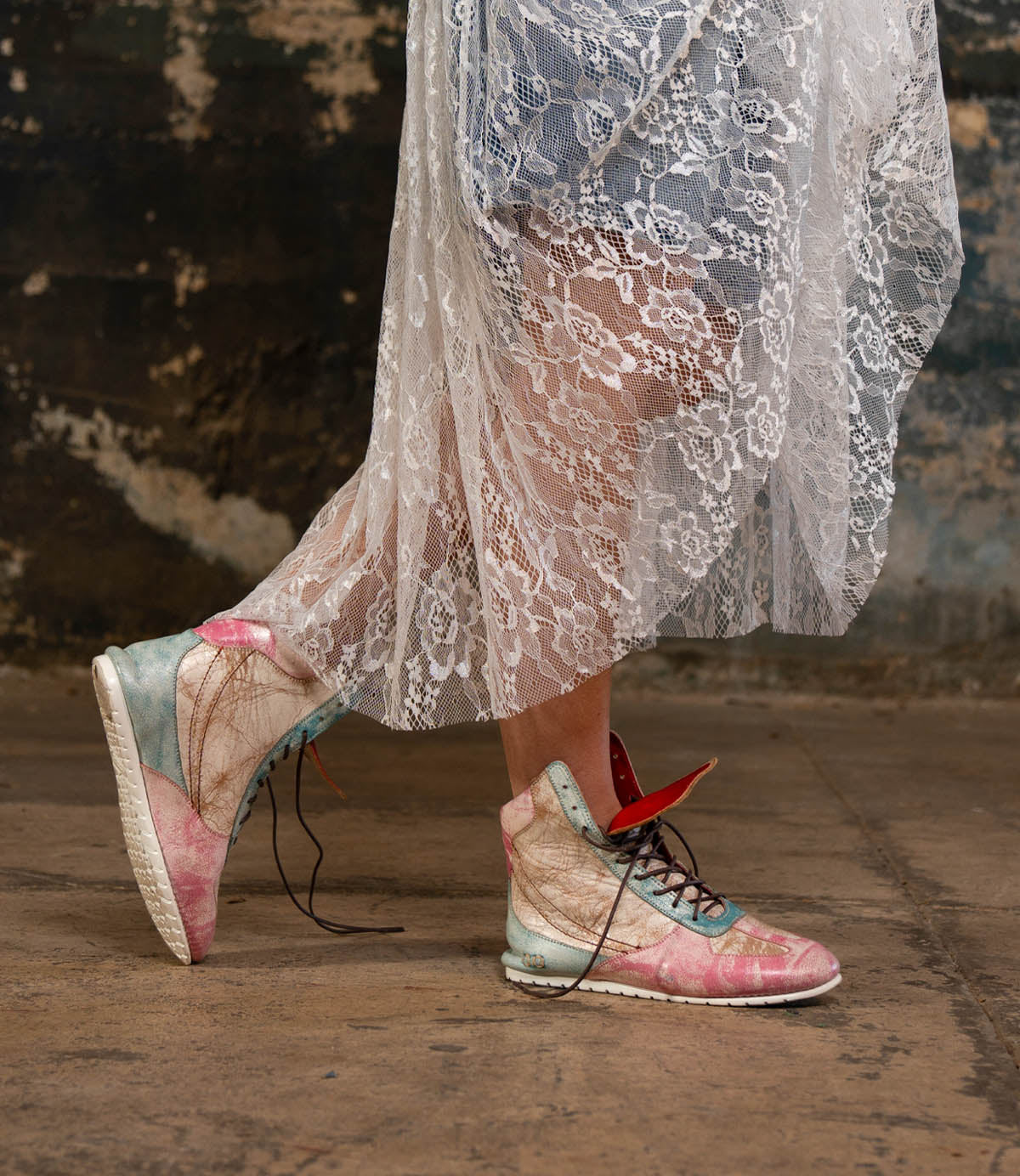 A person wearing the Bed|Stü Breath pastel-colored high-top statement sneakers and a sheer white lace dress stands on a worn concrete floor, with a textured, weathered wall in the background.