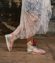 A person wearing the Bed|Stü Breath pastel-colored high-top statement sneakers and a sheer white lace dress stands on a worn concrete floor, with a textured, weathered wall in the background.