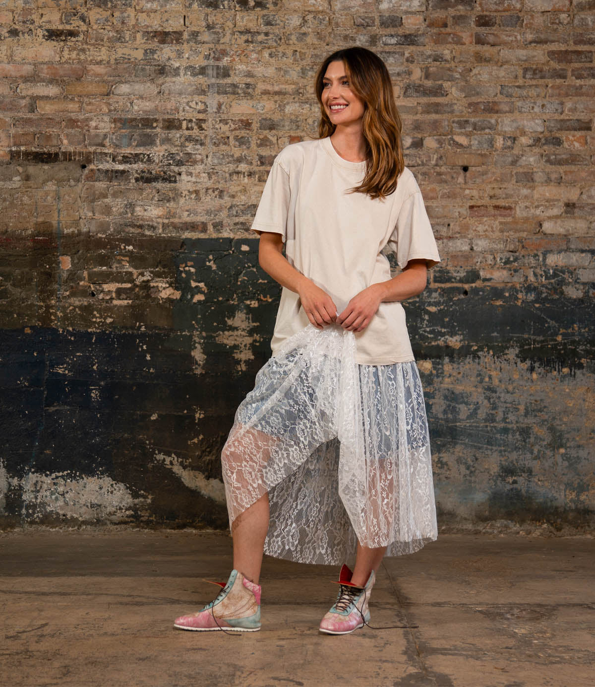 A woman stands indoors against a brick wall, smiling as she holds her skirt, dressed in a beige t-shirt, sheer lace skirt layered over shorts, and statement sneakers—the Breath sneakers by Bed|Stü.